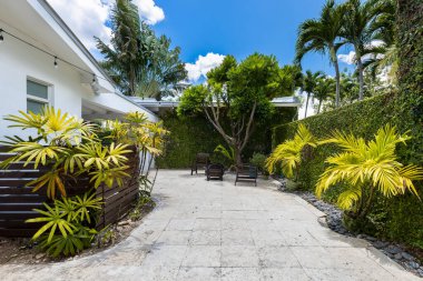 Interior patio of mansion with walls with vines, tropical plants, outdoor chairs, fire pit, tile and stone floor, string of hanging lamps, palms and blue sky