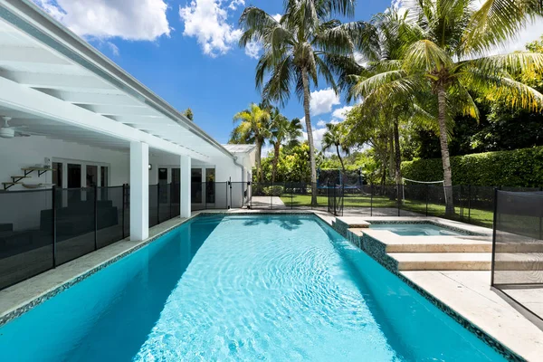 Backyard of home with fenced pool and spa, park with short grass, veranda with outdoor furniture, palms, blue sky
