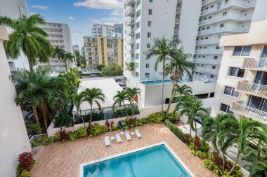 Point of view of swimming pool in the West bay building, in Miami, with red brick floor, tropical plants around, balconies, umbrellas with sun loungers, blue sky
