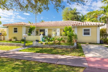 Beautiful yellow house facade in the La Gorce neighborhood of Miami Beach, classic style, white columns, short grass, potted plants, tropical vegetation, path to the front door, red tiles and blue sky