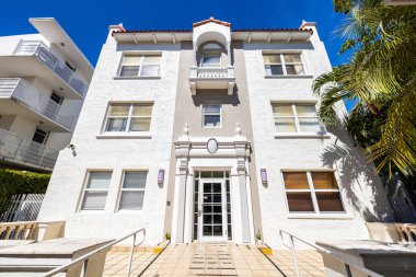 White and beige color colonial style building entrance, located in South Beach, Miami Beach, USA, palms tropical plants around, tiled floor with ramp, red tiles, surrounded by buildings, and blue sky