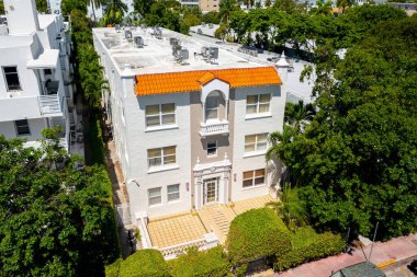 White and beige color colonial style building entrance, located in South Beach, Miami Beach, USA, palms tropical plants around, tiled floor with ramp, red tiles, surrounded by buildings, and blue sky