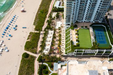 Miami Plajı 'ndaki Kuzey Sahili' nin insansız hava aracı görüntüsü, Akoya olarak bilinen bina, turkuaz deniz, şemsiyeler, modern binalar ve kuleler, mavi gökyüzü.