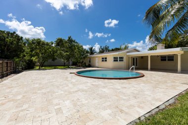 Miami 'de, çevresi yeşillik, dairesel havuz, veranda, ağaçlar, palmiye ağaçları, bitkilerle kaplı çiçek yatakları, çimen, çimento zemin ve mavi gökyüzü ile dolu çağdaş tarzda güzel bir evin verandası.