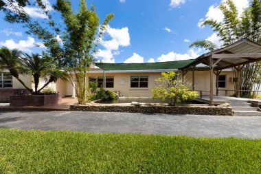 Miami 'deki çağdaş bir evin girişi, etrafında büyük miktarda yeşillik, yapay bir gölet, ağaçlar, palmiye ağaçları, bitkilerin olduğu çiçek yatakları, kısmi gölgeler ve mavi gökyüzü.