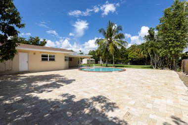 Miami 'de, çevresi yeşillik, dairesel havuz, veranda, ağaçlar, palmiye ağaçları, bitkilerle kaplı çiçek yatakları, çimen, çimento zemin ve mavi gökyüzü ile dolu çağdaş tarzda güzel bir evin verandası.