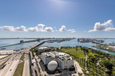 Miami şehir merkezindeki ticari alanın arkasından havadan çekilen görüntüler parklar, köprüler, arabalar, kahverengi binalar, müzeler, kuleler, rıhtımlar ve mavi gökyüzü.