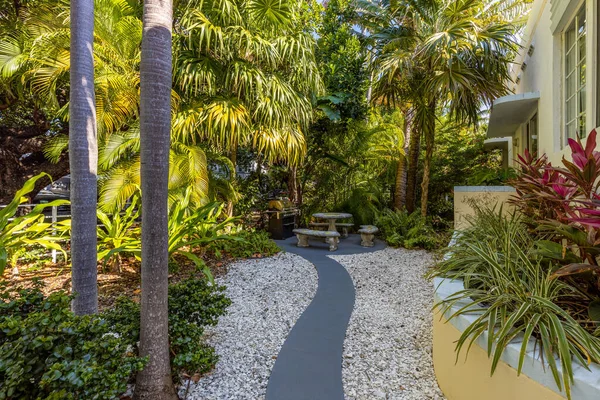 Beautiful backyard surrounded by tropical plants, outdoor grill, concrete table and chairs, flower beds with silver, palm trees, trees, paths and floor with decorative stones