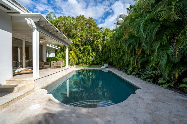 Beautiful backyard with concrete floor and pool, surrounded by tropical plants, porch with sun loungers, outdoor furniture, vines, located in Coral Gables