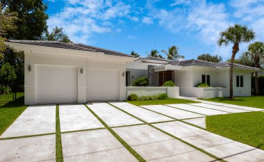 Zarif ve modern ev cephesi, Glenvar Heights, Miami-Dade 'de, beyaz duvarlar, siyah fayanslar, pencereli ahşap kapı, kısa çim, garaj yolu, ağaçlar ve palmiyeler, arka planda mavi gökyüzü.
