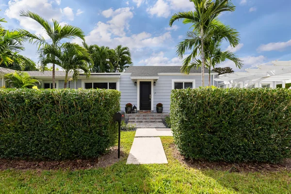 Facade of an elegant and modern wooden house in the West Flagler neighborhood, in Little Havana, driveway, short grass, privet walls, path to the front door, trees and palm trees, streets, light blue walls and blue sky