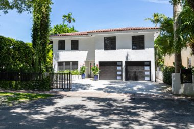 Coral Gables, Miami, FL, ABD 'deki Granada mahallesinde bulunan modern ve zarif beyaz bir evin girişi, çifte garaj yolu, çevresindeki harika bitki örtüsü, kırmızı fayanslar, mavi gökyüzü.