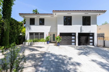 Coral Gables, Miami, FL, ABD 'deki Granada mahallesinde bulunan modern ve zarif beyaz bir evin girişi, çifte garaj yolu, çevresindeki harika bitki örtüsü, kırmızı fayanslar, mavi gökyüzü.