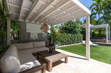 Coral Gables, Miami, FL, ABD 'deki Granada mahallesinde bulunan beyaz bir evin arka bahçesinde açık mobilyalar, kısa çimen alanı, açık mutfak, palmiyeler ve arka planda mavi gökyüzü olan güneşli şezlonglar yer alıyor.
