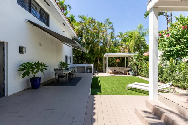 Backyard of a white house located in the Granada neighborhood in Coral Gables, Miami, FL, USA, covered patio with outdoor furniture, short grass area, outdoor kitchen, palms and sun loungers with blue sky in the background
