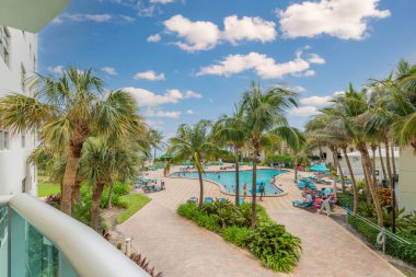 Hollywood South Central Beach 'teki bir otel havuzunun balkonundan bir sürü insan, şemsiyeler, şezlonglar, turkuaz deniz, palmiyeler, mavi gökyüzü.