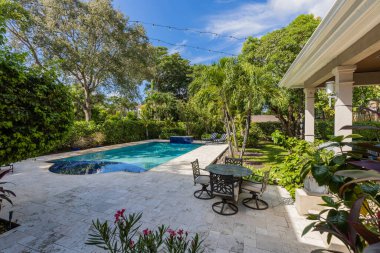 Zarif ve lüks malikanenin arka bahçesi, avuçlar ve bir sürü bitki, güzel havuz, kapalı teras, açık mobilya, kısa çim, basketbol sahası ve bulutlu mavi gökyüzü.