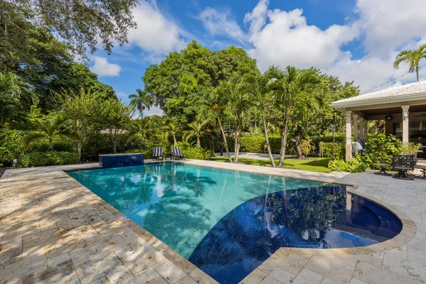 Backyard of elegant and luxurious mansion, with palms and lots of plants around, beautiful pool, covered patio, outdoor furniture, shortgrass, basketball court and blue sky with clouds