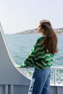 Kazaklı esmer bir kadın feribotta dikiliyor. Arka planda deniz ve Prenses adaları var.
