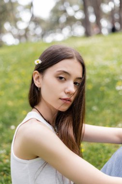 Portrait of brunette woman in top with daisy in hair looking at camera in park  clipart