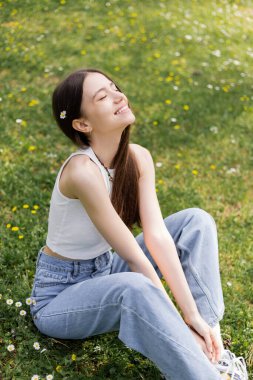 Carefree young woman in jeans and top sitting on lawn in park 