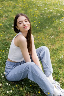Overjoyed woman in top and jeans looking at camera while sitting on lawn with flowers in park  clipart