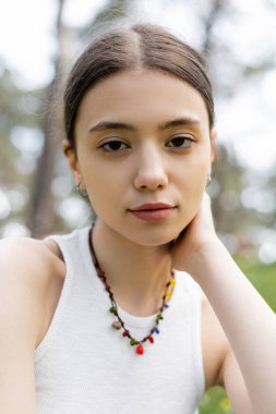 Portrait of young brunette woman in top and necklace looking at camera outdoors  clipart