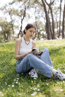 Genç bir kadın parkta çiçeklerle çayırda otururken kitap okuyor. 