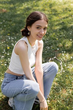 Tepedeki kaygısız genç kadın ve parktaki çiçeklerle çayırdaki kameraya bakan kot pantolon. 