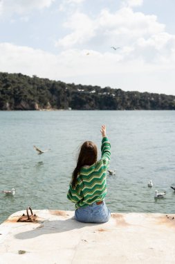 Back view of young woman sitting on pier near blurred seagulls on water in Turkey  clipart