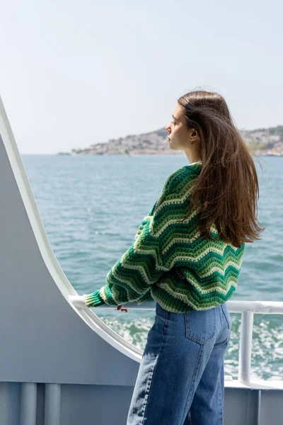Kazaklı esmer bir kadın feribotta dikiliyor. Arka planda deniz ve Prenses adaları var.