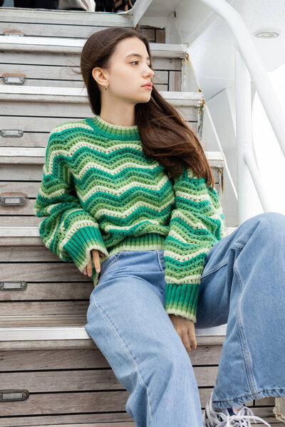 Young brunette woman in sweater and jeans sitting on stairs on yacht 