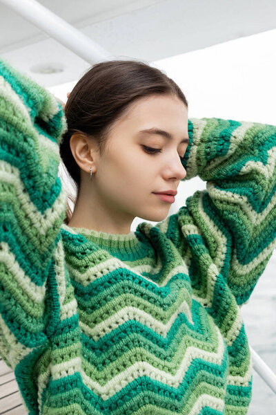 Pretty young woman in knitted sweater touching hair on yacht 