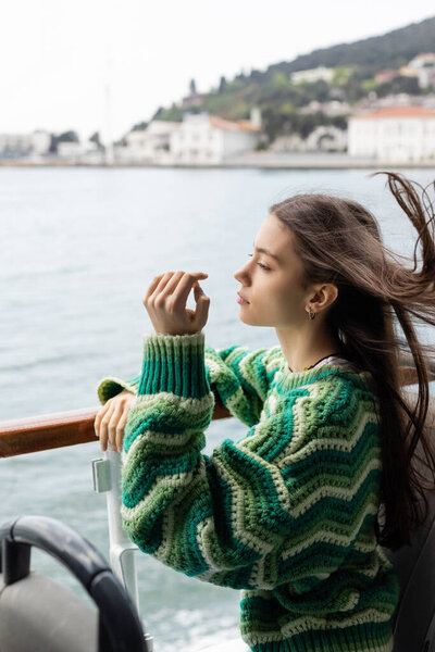 Side view of young woman in sweater standing near railing on yacht in Turkey 