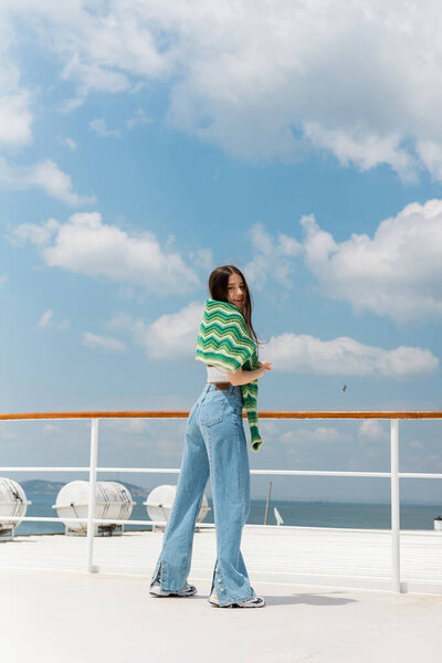 Smiling woman in jeans and sweater looking at camera while standing on yacht in Turkey 