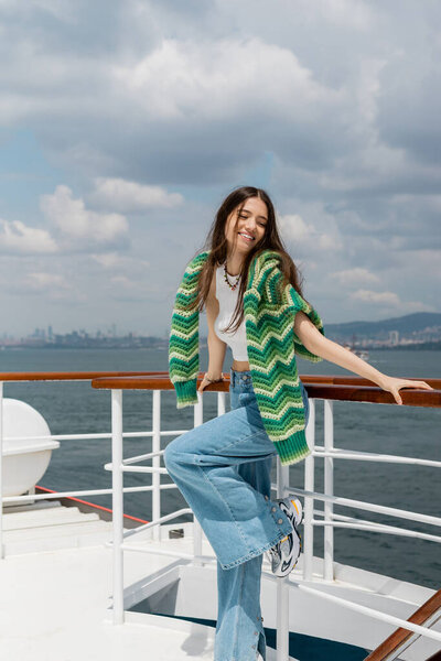 Позитивная молодая женщина в трикотажном свитере и джинсах, стоящая рядом с перилами яхты в Турции 