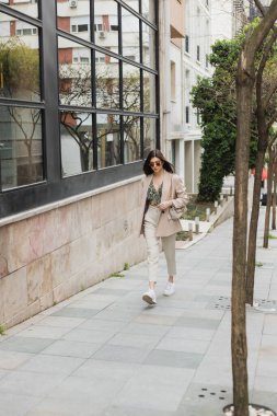 Şık güneş gözlüklü, şık giyinen, beyaz pantolonlu, bej ceketli, ağaçların yanında el çantalı ve İstanbul 'da modern binalı genç esmer kadın. 