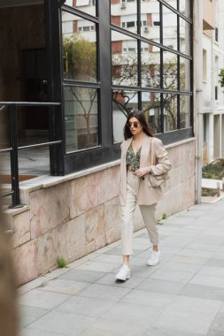 Şık güneş gözlüklü, modaya uygun giyinen, beyaz pantolonlu, bej ceketli, modern binanın yanında elinde çantasıyla gezen İstanbul 'da camları olan esmer bir kadın. 