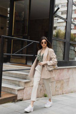 Şık güneş gözlüklü, modaya uygun giyinen beyaz pantolonlu, bej ceketli, modern binanın yanında el çantalı, pencereli ve merdivenli genç bir kadın İstanbul caddesinde yürüyor. 