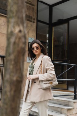 Şık güneş gözlüklü, modaya uygun giyinen, beyaz pantolonlu, bej ceketli, modern binanın yanında el çantasıyla yürüyen esmer ve İstanbul caddesinde bulanık ağaç gövdesi olan genç bir kadın. 