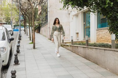 Uzun saçlı, bej pantolonlu, kırpılmış bluz ve zincir kayışlı el çantalı genç kadın modern binanın yanında yürüyor, park edilmiş arabalar ve İstanbul 'da yeşil ağaçlar,