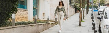 Çağdaş binanın, arabaların ve yeşil ağaçların yanında yürüyen bej pantolonlu, kırpılmış bluz ve zincirli çantalı genç kadın İstanbul 'da kentsel cadde, turist, afiş 