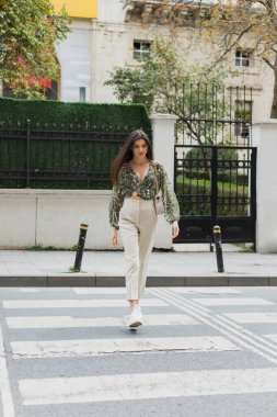 chic woman with brunette long hair in trendy outfit with beige pants, cropped blouse and handbag with chain strap walking on crosswalk of urban street in Istanbul, blurred house on background  clipart