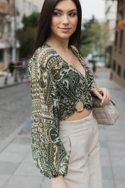 Şık, esmer, uzun saçlı, modaya uygun giyimli, bej pantolonlu, kırpılmış bluzlu ve zincir kayışlı el çantalı bir kadın İstanbul 'da sokak ortasında ayakta duruyor ve gülümsüyor. 