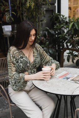 young woman with long brunette hair and makeup sitting on chair near round bistro table with newspaper and holding paper cup with coffee near blurred plants on terrace of cafe in Istanbul  clipart
