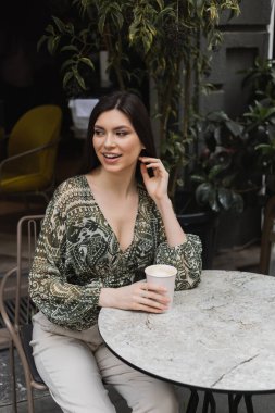 cheerful woman with long brunette hair and makeup sitting on chair near round bistro table and holding paper cup with coffee near blurred plants on terrace of cafe in Istanbul  clipart