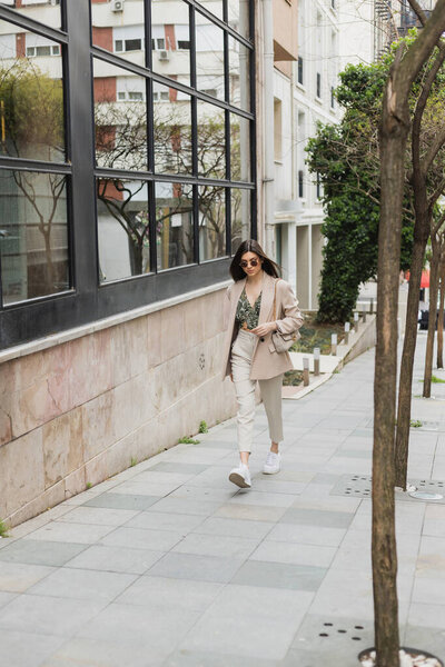 young brunette woman in stylish sunglasses and trendy outfit with white pants and beige blazer walking with handbag near trees and modern building on street in Istanbul 