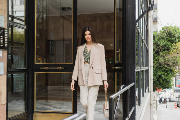 brunette and young woman in trendy outfit with white pants, blouse and beige blazer walking out of modern building while holding handbag with chain strap on street in Istanbul 