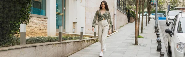 stock image young woman with long hair in beige pants, cropped blouse and handbag with chain strap walking near modern building, cars and green trees on urban street in Istanbul, tourist, banner 
