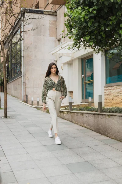 young woman with long hair in trendy outfit with beige pants, cropped blouse and handbag with chain strap walking with hand in pocket near modern building and green tree on street in Istanbul